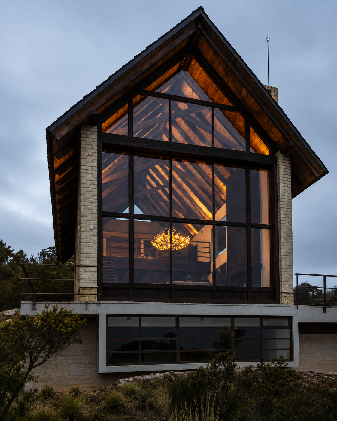 Fachada de vivienda unifamiliar al atardecer con un gran ventanal a dos aguas que revela la estructura de madera y una lámpara cálida en el interior, sobre base de ladrillo y piedra — proyecto GAT 02 de LINEA PRIMA, arquitectos en Bogotá.